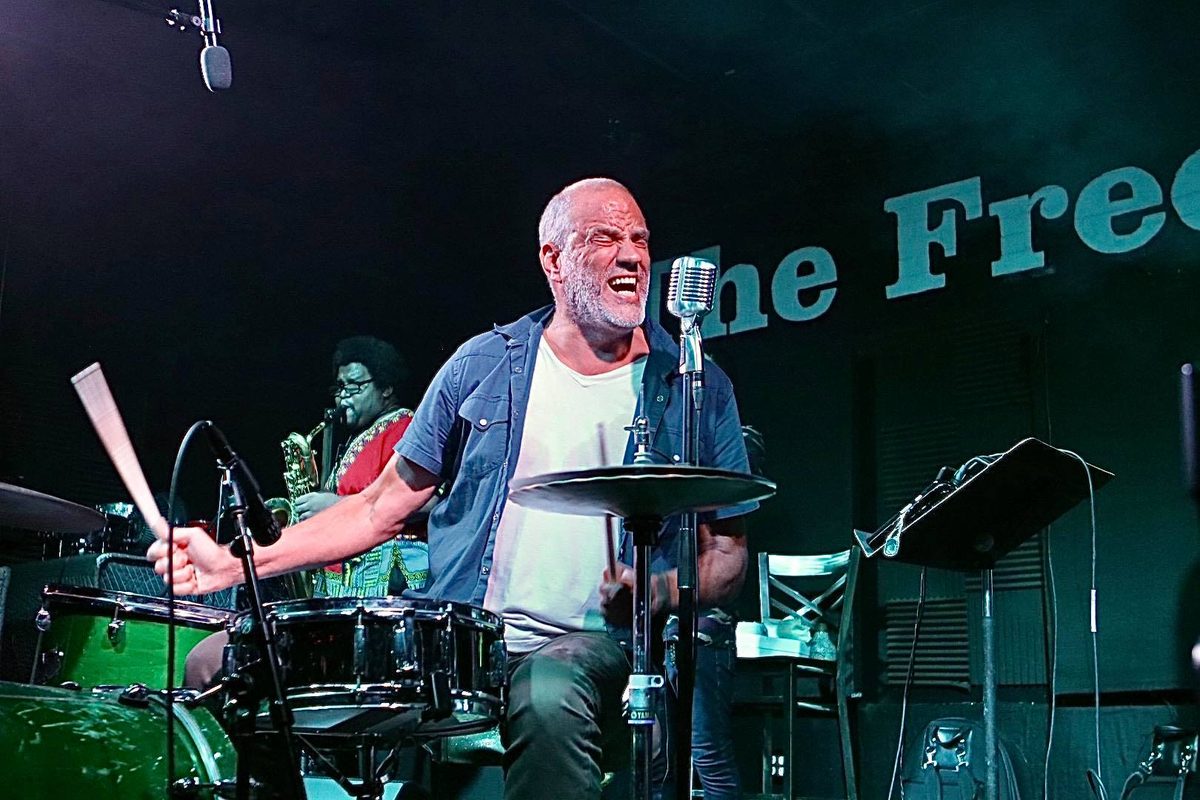 John Jay Myers drumming and singing on The Free Man stage, with sax behind him